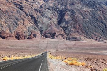 Endless highway and cars at the foot of the huge red and black rocks in Death Valley