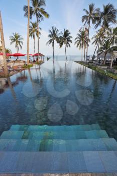 Marble steps leave under pool water. A beach on coast of the Thai gulf, red beach umbrellas, plank beds and palm trees against the sea