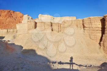 The ancient crumbling mountains of various forms in the Judean desert. The giant shadow of the photographer with a photo bag on the bottom of the parched canyon