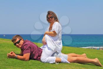 Happy young couple relaxing on the grass by the sea on a summer day. Charming young woman is pregnant