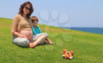 Beautiful young pregnant woman rests with lovely little son on the grass near the sea