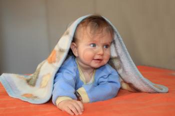 The charming  blonde baby with the blue eyes, covered by a blanket, joyfully smiles