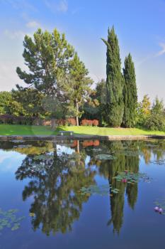 Smooth water of a pond reflects coastal cypresses and picturesque bushes. Magnificent Italian park at sunset