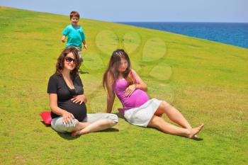 Two young pregnant woman in sunglasses posing on the grass lawn. Young son of one of them comes up to them