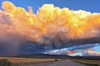 Summer rain. The huge cumulonimbus cloud is shined with the sunset sun. Rain streams shine orange light. The cloud hangs over the gravel road