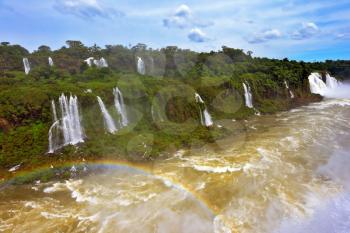 Raging and roaring water in the waterfalls of Iguazu. Turbid yellow-brown waves flow down. Above the water is a picturesque rainbow