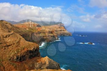 Eastern tip of the island of Madeira. Rocks steeply in the blue waters of the Atlantic Ocean.  Over a cliff on the ocean breeze are the windmills
