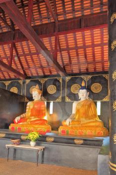 Two sitting next to the statue of the Buddha under a canopy. The statues dressed in gilded clothing