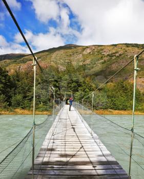 Suspension bridge across mountain river. In the middle of the bridge woman - tourist photographs the raging torrent