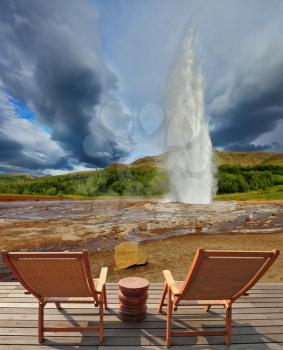 Pillar of hot water and steam from forcing its way out of the ground.  Geyser Strokkur in Iceland. Two lounge chairs and  small table on  wooden platform for easy observation