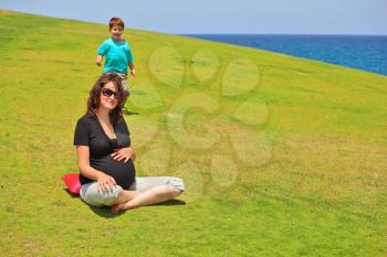 Young pregnant woman with sunglasses posing for a photograph on the grass lawn. Her young son laughing runs to her
