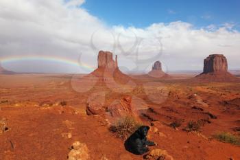 A large black dog under a rainbow in the red desert. The famous Mittens in Monument Valley after the rain
