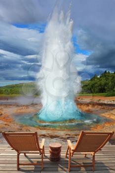 Pillar of hot water and steam from forcing its way out of the ground. Two lounge chairs and  small table on  wooden platform for easy observation.  Geyser Strokkur in Iceland