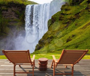Magnificent famous waterfall Skógafoss, Iceland. A powerful jet Skógar river falls from  large glacier. On stony ground in front of the waterfall are two wooden deck chairs