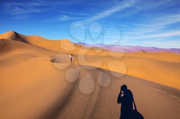Orange sand dunes in Death Valley, California. A middle-aged woman in striped T-shirt carries a camera and tripod