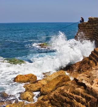  The fisherman sits with a fishing tackle at the sea on top  an ancient stone wall
