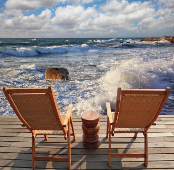 Charming lookout for two. Two convenient comfortable guest sun loungers and a bedside table are worth on a wooden platform. It is well-admired stormy sea surf