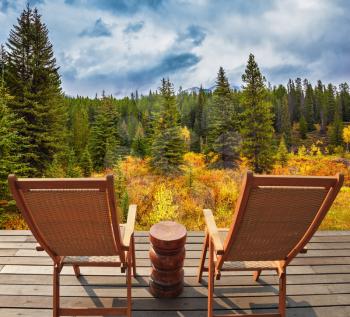 On the wooden platform there are two wicker deck chairs. The lush colorful autumn in the Rocky Mountains of Canada