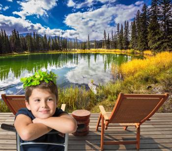 The charming boy sits on the shore of a small circular lake. The  autumn grass and trees. Rocky Mountains of Canada 