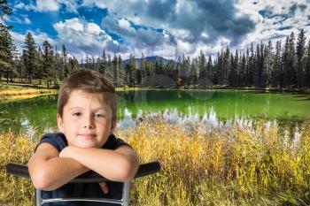 Handsome boy sitting on the shore of small lake. The yellow and orange autumn grass and trees. Rocky Mountains of Canada 