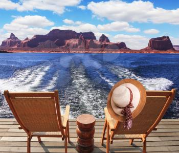 At the stern of the vessel are two deck chairs. On the back of one hanging elegant ladies straw hat. Waves from the boat cut through the Lake Powell on the Colorado River