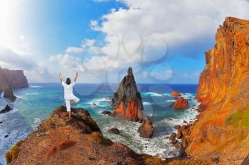  The woman ashore in a white suit for yoga carries out a pose Tree. Atlantic storms. Colorful pinnacles lit sunset. Arid eastern tip of the island of Madeira