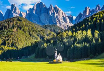  The symbol of the valley Val di Funes - church of Santa Maddalena. Rocky peaks and forested mountains surrounded by green Alpine meadows. Tirol, Dolomites. Sunny warm autumn day