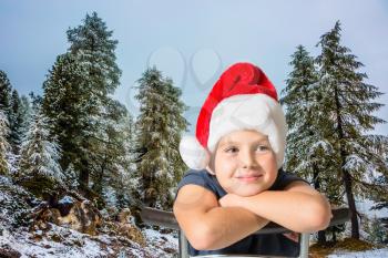 Charming eight-year boy in Santa Claus hat smiling.  Pine forest in mountains are covered with the first snow
