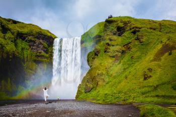 Grand waterfall with rainbow in the water mist Skogafoss. Elderly woman performs asana tree of hatha yoga before falling water