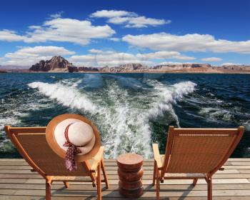 Waves from the boat cut through the Lake Powell on the Colorado River. At the stern of the vessel are two deck chairs. On the back of one hanging elegant ladies straw hat