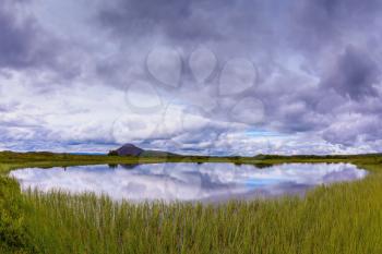  Small lake surrounded by green meadow. In the smooth water of cold lake reflects cloudy sky. Summer Iceland
