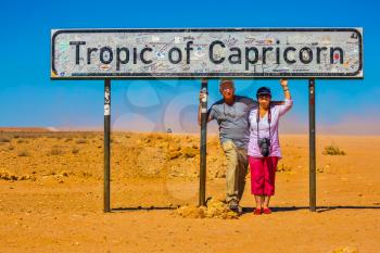 Happy elderly couple of tourists. South Tropic, or Tropic of Capricorn, - the famous road sign in Namibia. The concept of extreme and active tourism