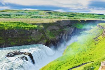 Grandiose Gullfoss in Iceland. In July raging water is shined with the bright morning sun. River banks grew with a green northern moss