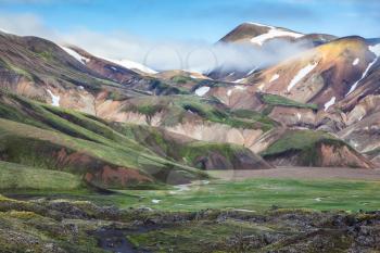 Snow and fog lies in the hollows of colorful rhyolite mountains.  Summer morning in the National Park Landmannalaugar, Iceland. Green Valley is flooded with melt water