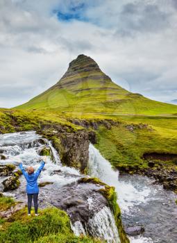 Middle-aged woman tourist admires the beauty of nature. Threaded full-flowing waterfall Kirkjufell Foss on the grassy mountains
