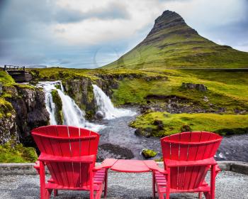 Red paired chaise lounges for rest. Summer in Iceland. The concept of exotic and ecological tourism. Cascading waterfall Kirkjoufellfoss at the foot of the mountain Kirkjoufell