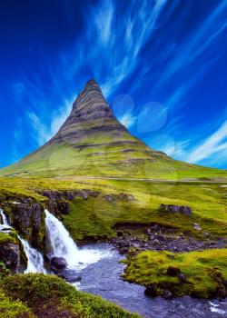 The famous mountain in Iceland is Kirkufell. At the foot of the mountain cascading waterfalls Kirkjoufellfoss. Cirrus clouds in the blue sky. The concept of exotic and extreme tourism