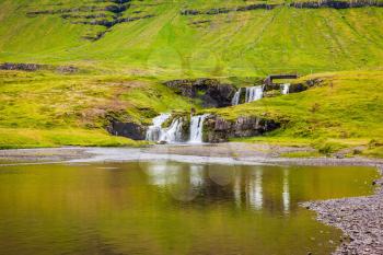 Cascading waterfall Kirkjoufellfoss is reflected in a large puddle at the foot of the mountain Kirkjoufell. Summer in Iceland. Concept of exotic and extreme tourism