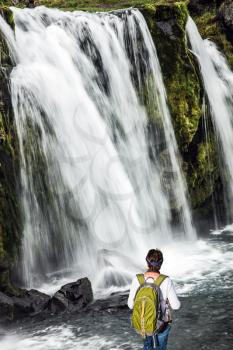 Elderly woman -  traveler with a large backpack admiring the powerful jets of the waterfall Kirkjoufellfoss. Summer in Iceland. Concept of exotic and extreme tourism 