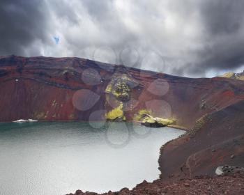Iceland in July. Oval blue lake in the crater of the volcano cooled down. The lake shores are made of red rhyolite