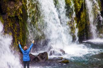 Concept of exotic and extreme tourism. Waterfall Kirkjoufellfoss. Elderly woman -  traveler enthusiastically raised her hands. Summer in Iceland