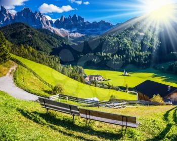 The bright morning sun illuminates the green valley, rural scenic farm and the church.  The concept of eco-tourism in Alpine meadows. Rural dirt road