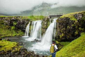 Cascading waterfall Kirkjoufellfoss. Summer in Iceland. Elderly woman -  traveler with a large backpack admiring the waterfall. Concept of exotic and extreme tourism 