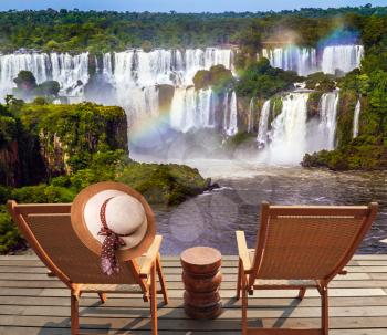  Incredible exotic waterfalls of Iguazu in South America. Two comfortable wooden chaise lounges face waterfalls. On one hangs an straw female hat. Concept of exotic and eco-tourism