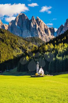  The symbol of the valley Val di Funes - church of Santa Maddalena. Tirol, Dolomites. Rocky peaks and forested mountains surrounded by green Alpine meadows. Sunny warm autumn day