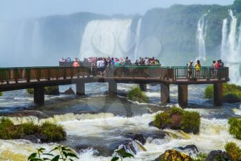 Hundreds of tourists watch the grandiose waterfalls of Iguazu from the bridge. The world of roaring water. Iguazu Falls in South America. Concept of active and extreme tourism