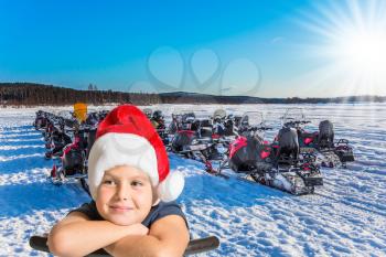 Soon Christmas. Concept of active winter tourism. Handsome boy in a red Santa Claus hat. Tourist train from snowmobiles moves along the frozen river