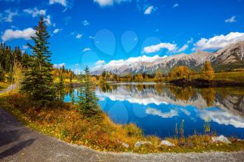  The concept of hiking. Path around the lake. Canmore, near Banff National Park. Mountains and scenic cumulus clouds are reflected in the water