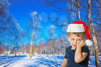  New Year in Lapland. Very handsome little boy in a red Santa Claus hat in winter snow-covered aspen grove. Sunny and snowy winter day. Concept of the children's tourism