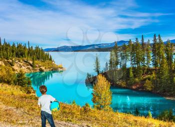 Nine-year-old boy in jeans with a globe in his hands admires the lake. Exquisite Abraham Lake with turquoise water. Indian Summer in the Rockies of Canada. Concept of ecological and active tourism